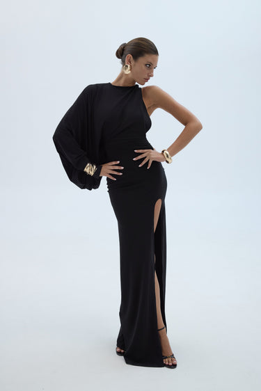 A woman modeling a floor-length black gown with a one-shoulder draped sleeve, a thigh-high slit, and gold jewelry against a light gray studio background.