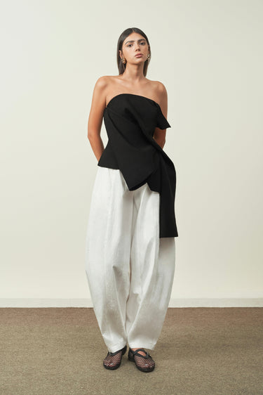 Woman wearing a black one-shoulder top and white pants against a plain background