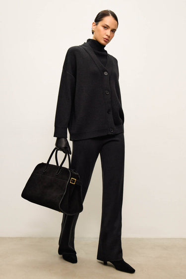 Woman in a black outfit holding a matching handbag against a plain background