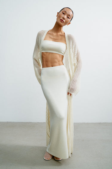 Woman wearing a white outfit with a beige cardigan against a plain background