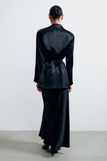 Person wearing a black outfit with a long skirt and jacket on a plain background