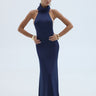 A woman modeling a sleeveless navy blue maxi dress with a high mock neck and cowl tie detail, posed against a light gray studio background.