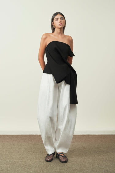 Woman wearing a black one-shoulder top and white pants against a plain background