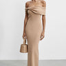 Woman wearing a beige off-shoulder dress holding a matching handbag against a white background