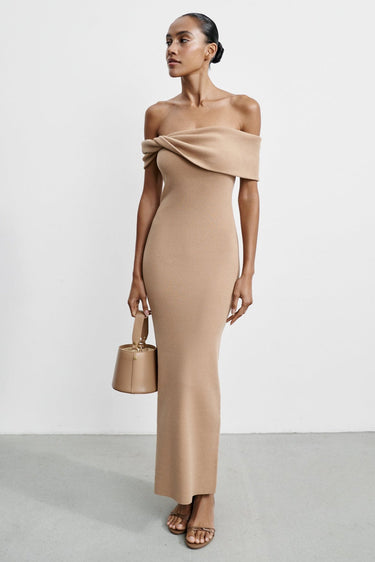 Woman wearing a beige off-shoulder dress holding a matching handbag against a white background