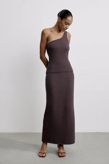 Woman wearing a long, dark purple one-shoulder dress against a plain background