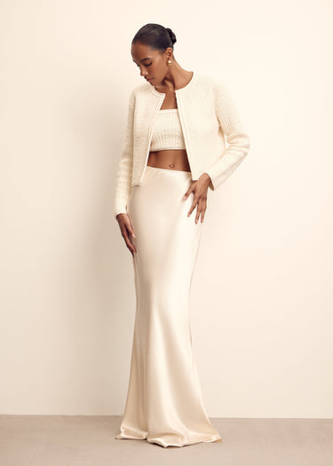 Woman wearing a white outfit with a long skirt and cropped jacket against a beige background