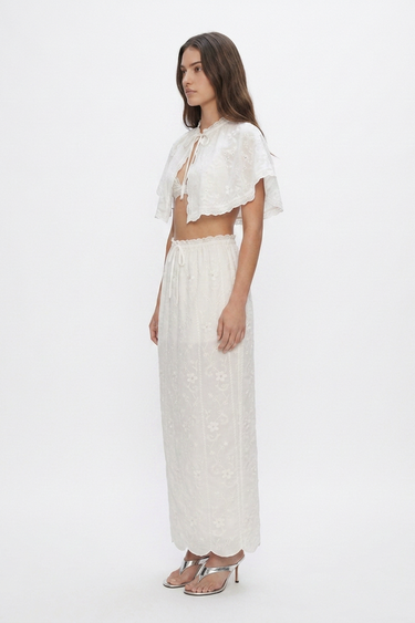 A woman modeling a white two-piece outfit consisting of a short-sleeve cropped lace top with ruffled edges and matching high-waisted maxi skirt, photographed against a plain white studio background.